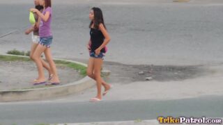 Two young women walking on the sidewalk, one wearing a purple shirt and the other wearing a black shirt, both in shorts and flip-flops